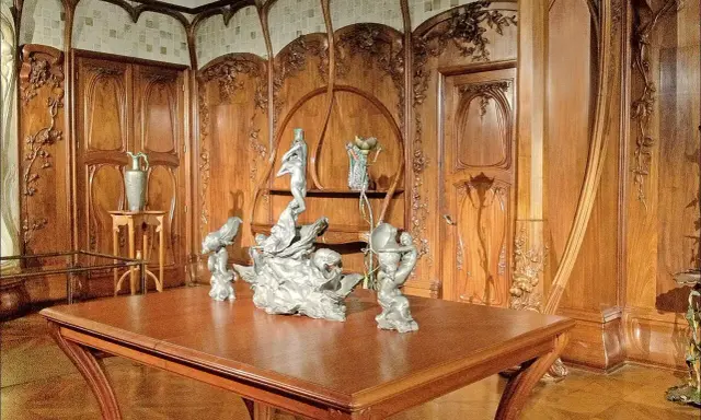 Intérieur Art Nouveau orné de boiseries sculptées, avec une table en bois et des sculptures, évoquant l'œuvre d'Alexandre Charpentier.