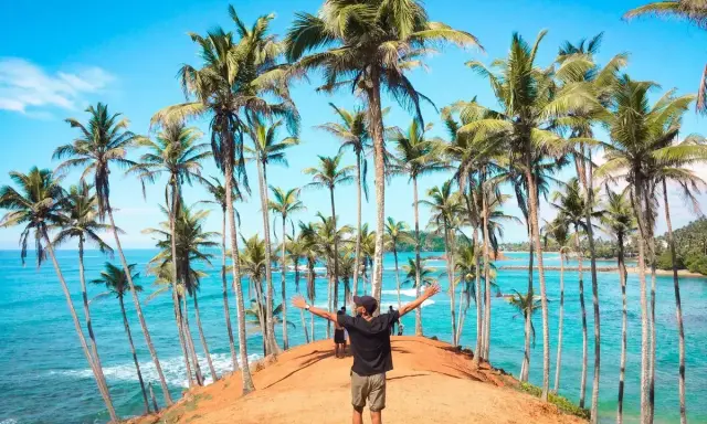 Człowiek z rozłożonymi rękami na tle palm i turkusowego oceanu. Marzysz o podróży? Sri Lanka ile trwa lot, warto sprawdzić!