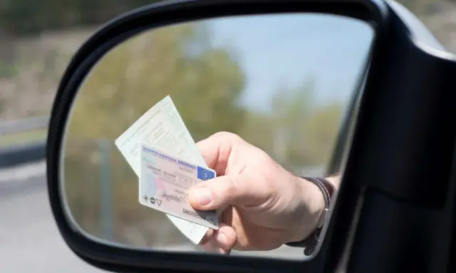 Mano sujetando un carnet de conducir y un documento de identidad reflejados en el espejo retrovisor de un coche.