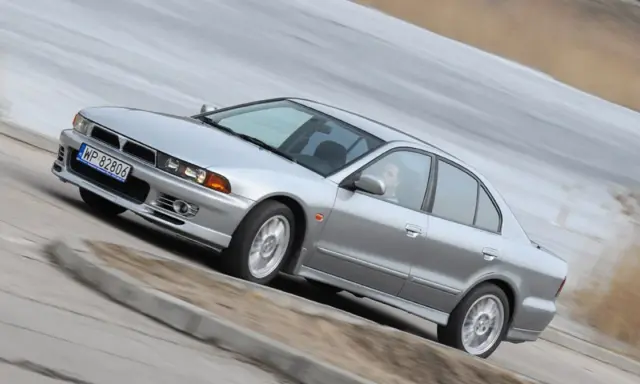 Srebrny Mitsubishi Galant z gracją pokonuje zakręt. Kierowca w okularach pewnie prowadzi auto.