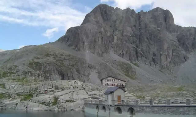 Rifugio sul lago della Vacca, con imponente parete rocciosa alle spalle.