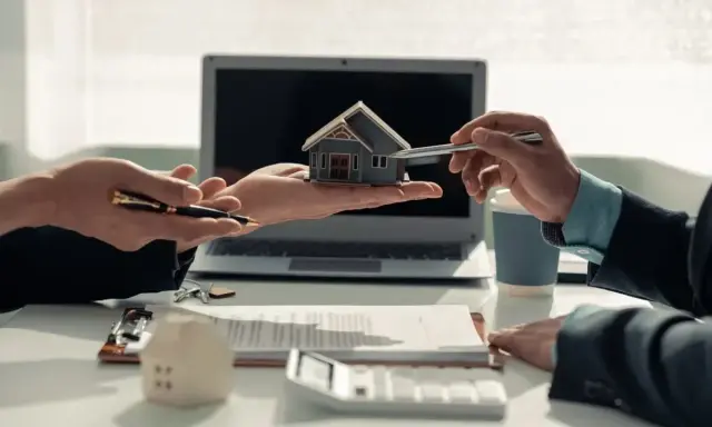 Deux mains présentent une maquette de maison, symbolisant comment devenir investisseur immobilier. Un contrat et un ordinateur sont visibles.