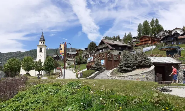 A picturesque village in Chamois, Italy, with a church tower, wooden chalets, and lush greenery. Scopri cosa vedere in questo incantevole borgo alpino.