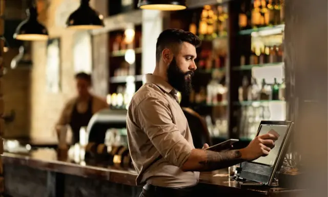Hombre con barba gestionando un bar, usando una tablet y una pantalla táctil para procesar pagos.
