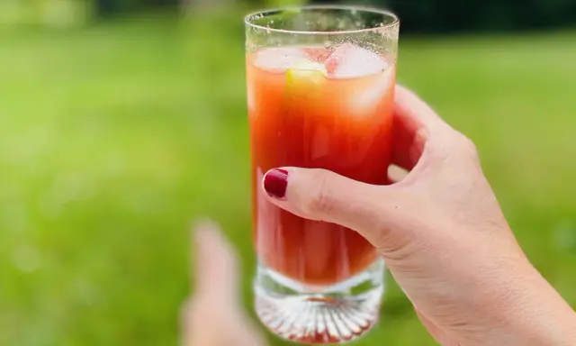 A hand with red nail polish holds a tall glass of Bloody Mary with ice and a cucumber slice, outdoors.
