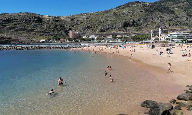 Sonniger Strand in Machico, Madeira, mit vielen Menschen, die im klaren Wasser planschen und sich am Sandstrand sonnen.
