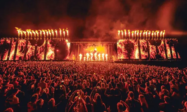 Un'immensa folla partecipa a un festival tekno, illuminata da fiammate spettacolari e schermi con immagini di fuoco.