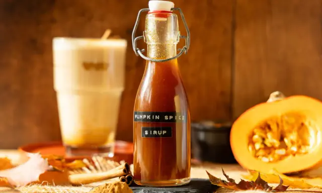 Flasche mit goldbraunem Pumpkin Spice Sirup, daneben ein Latte Macchiato, Kürbishälften und Herbstlaub.