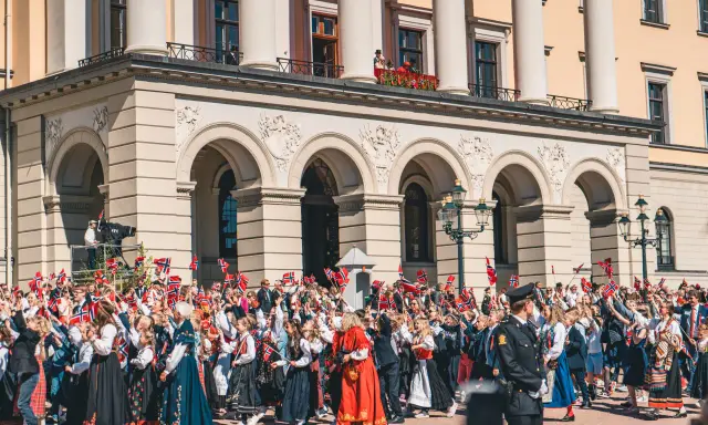 Tłum ludzi, w tym dzieci, świętuje, machając norweskimi flagami. Wiele osób nosi tradycyjny strój norweski, ozdobiony haftami i kolorowymi wzorami.