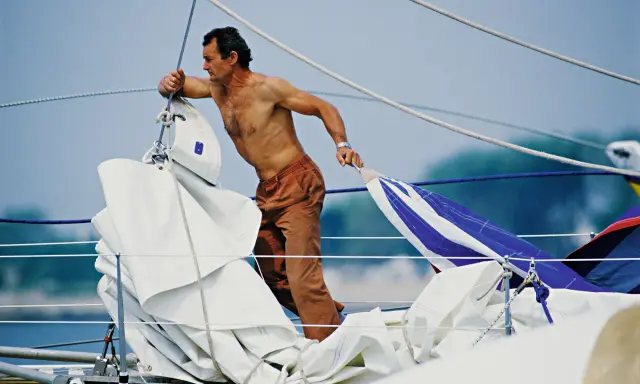 Un marinier, peut-être Eric Tabarly, manipule des voiles sur un bateau. Sa fortune est dans le vent et l'océan.