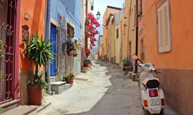 Un vicolo colorato con case arancioni e blu, fiori rossi e una Vespa bianca. Perfetto per le tue idee weekend nord Italia.