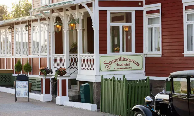 Strandflickornas Havshotell i Lysekil, en charmig röd byggnad med vita detaljer, välkomnar besökare. En klassisk bil står parkerad framför.