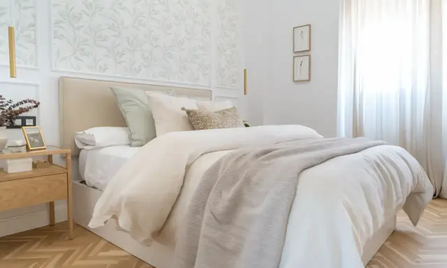 Dormitorios con papel pintado de hojas verdes en el cabecero. Cama con ropa de cama blanca y manta gris. Mesita de noche de madera.