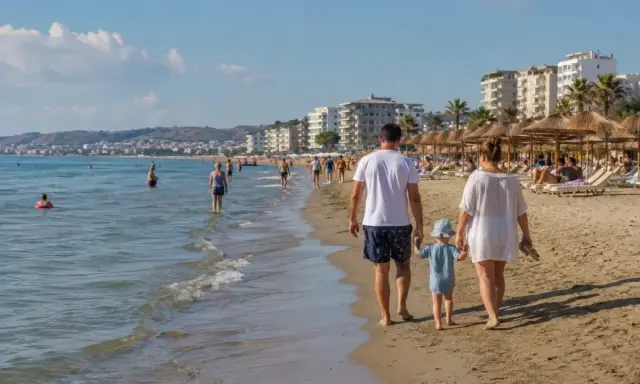 Rodzina spaceruje po piaszczystej plaży w Durrës. W tle widać morze, budynki i parasole plażowe.
