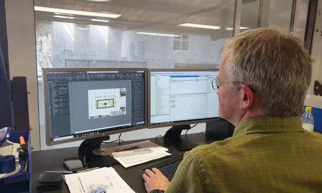 Mann am Computer, der Baupläne nach DIN 4426 erstellt. Auf dem Bildschirm sind CAD-Zeichnungen und eine Liste von Bauteilen zu sehen.