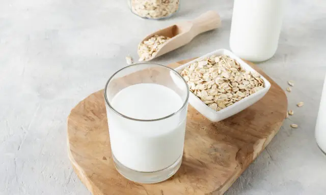 Ein Glas cremiger Hafermilch steht auf einem Holzbrett, daneben Schüsseln mit Haferflocken. Hafermilch gesund und lecker!