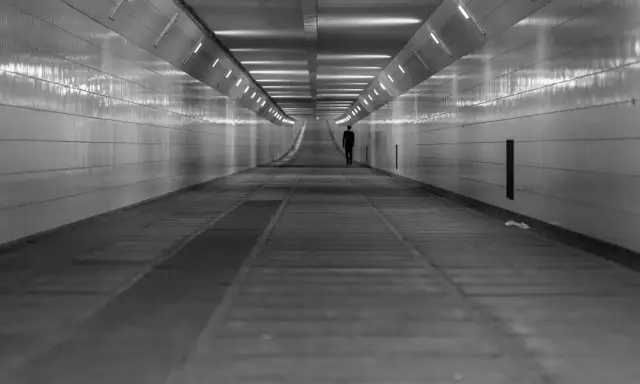 Człowiek w garniturze idzie przez długi, pusty tunel. Światła na ścianach tworzą wrażenie nieskończoności, jakby wchodził w szarą strefę.