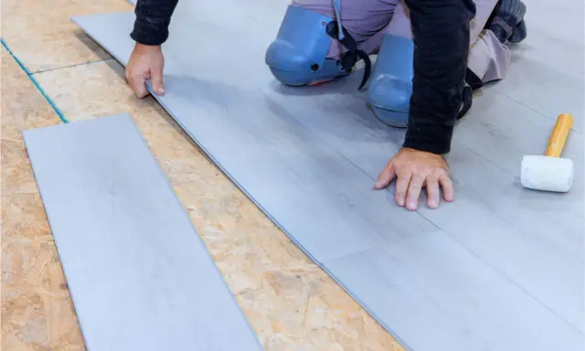 Man con rodilleras coloca cuidadosamente tablones de suelo laminado gris sobre una base de madera.