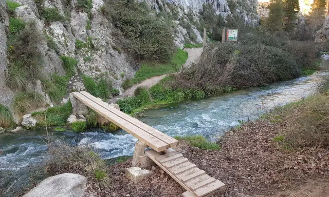 Puente de madera sobre un río caudaloso, parte de la ruta del zarzalar. Senderos y vegetación agreste flanquean el camino.