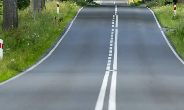 Droga wijąca się wśród zieleni, z podwójną białą **linią ciągłą** pośrodku.