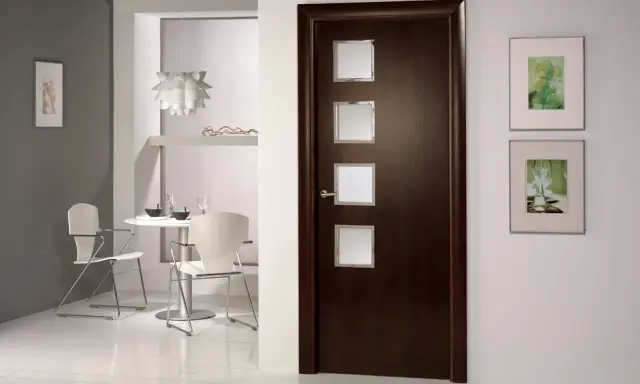 Puerta wengue con marcos de cristal. Paredes claras y un rincón de comedor moderno.