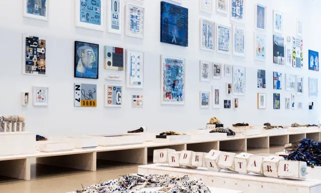 Konstutställning med verk av Carouschka Streijffert. Tavlor med grafiska motiv och text hänger på väggen. Framför står utställningsbänkar med konstobjekt.