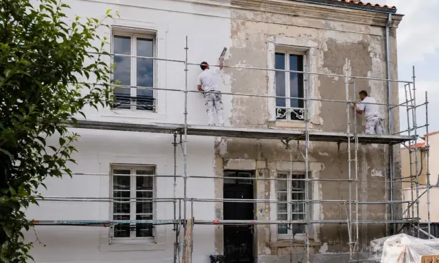 Deux ouvriers sur des échafaudages sont en train de refaire sa façade, appliquant une nouvelle couche de peinture blanche sur un mur décrépi.