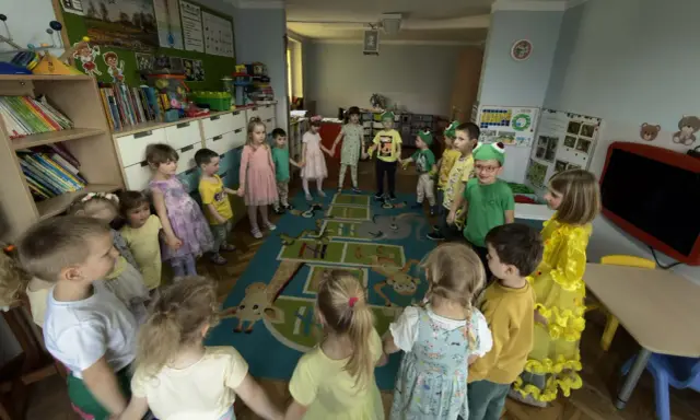 Dzieci trzymają się za ręce, tworząc krąg na dywanie z zabawnymi zwierzątkami. To radosna zabawa w **zespole szkolno przedszkolnym**.