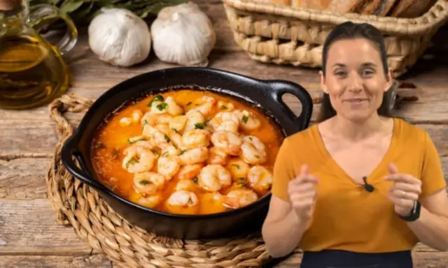 Una mujer sonríe mientras muestra cómo hacer gambas al ajillo, con un plato humeante y los ingredientes listos.