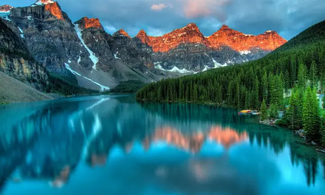 Turkusowe jezioro odbija ośnieżone szczyty gór o zachodzie słońca. W tle gęsty las iglasty. Park narodowy Banff zachwyca.