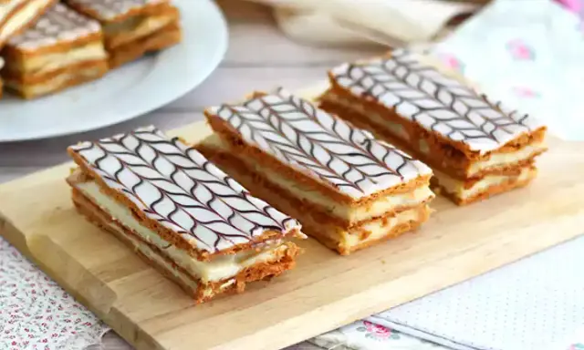 Tre läckra millefeuille-bakelser med lager av smördeg och kräm, toppade med vit glasyr och chokladmönster.