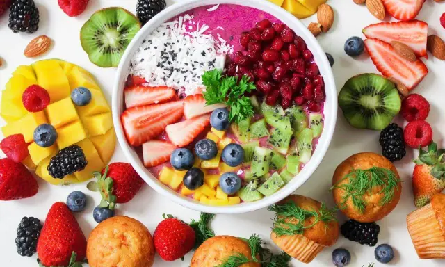 Kolorowa miska smoothie z owocami, jagodami, granatem i wiórkami kokosowymi. Obok mango, truskawki, kiwi, maliny i muffinki. Idealne owoce na noc.