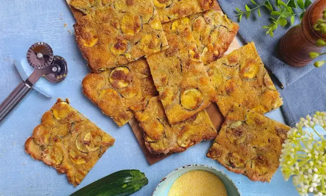 Fette dorate di schiacciata di zucchine, condita con erbe aromatiche, su un tagliere. Accanto, una zucchina fresca, farina di mais e un rotella taglia pizza.