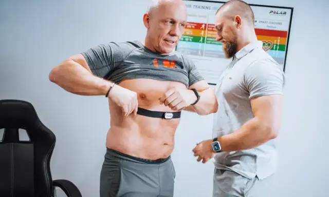 Mann legt Brustgurt für Muskelerhalt Training an, Trainer hilft.