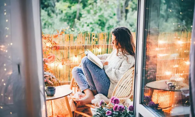 Przytulny jesienny balkon. Dziewczyna w swetrze czyta książkę, otoczona jesiennymi liśćmi i ciepłym światłem lampek.