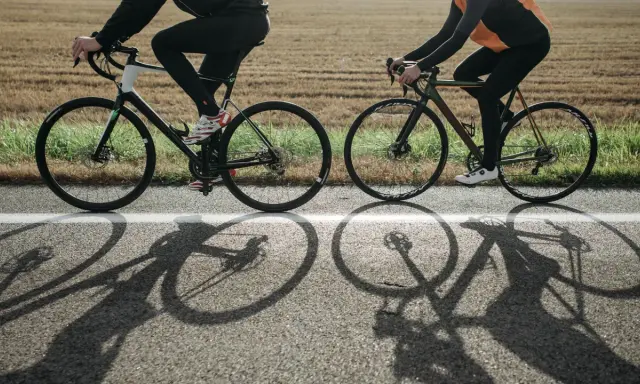 Dwie osoby na rowerach, w kaskach, jadące po drodze. Długie cienie rowerów na asfalcie.