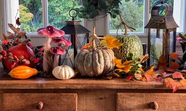 Décoration automne facile à faire : citrouilles, lanternes, fausses champignons et feuilles d'automne sur un buffet en bois devant une fenêtre.