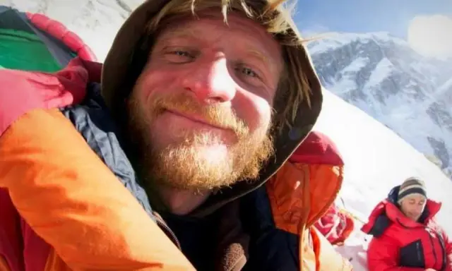 Uśmiechnięty alpinista w ciepłej kurtce, z brodą, na tle ośnieżonych gór. Może to być jego lek na chorobę wysokościową.