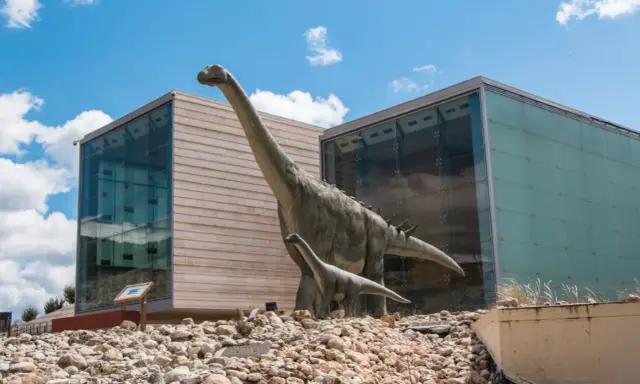 Dos dinosaurios gigantes, como los que verían los niños en la cuenca, posan frente a edificios modernos.