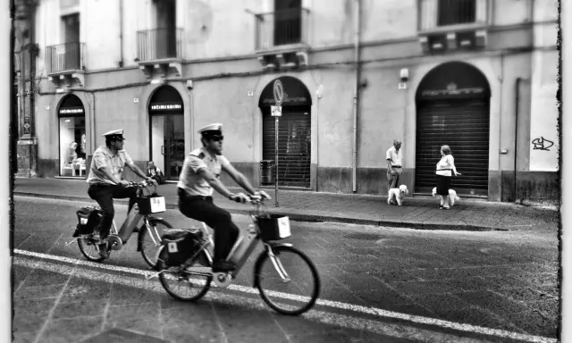 Due agenti in bicicletta pattugliano una strada italiana. Sul marciapiede, una coppia passeggia con i loro cani. Street photography Italia.