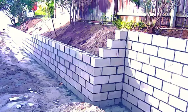 Un muro de contención de bloques de hormigón se está construyendo en un jardín, con tierra y vegetación detrás.