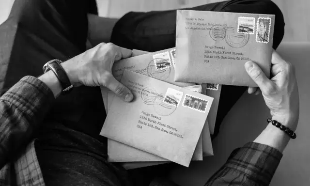 Dłonie trzymają stertę kopert, jakby przygotowywano korespondencję do osoby zmarłej. Na kopertach widnieją adresy i znaczki pocztowe.