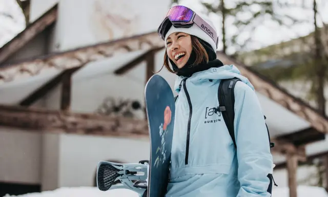 Uśmiechnięta snowboardzistka w jasnoniebieskim anoraku i goglach Anon. Gotowa na stok, trzyma deskę Burton.