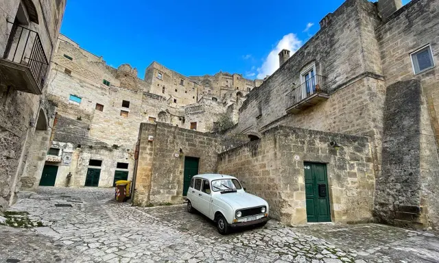 Un'auto d'epoca bianca parcheggiata tra le antiche case scavate nella roccia dei Sassi di Matera, sotto un cielo azzurro.