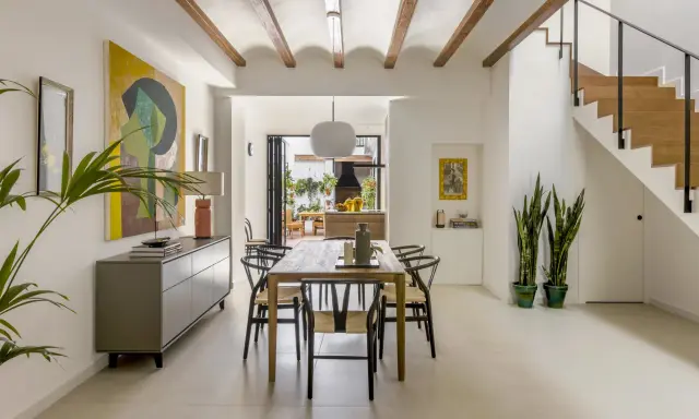 Comedor moderno en una casa entre medianeras, con vigas de madera en el techo, mesa de madera, sillas negras y una escalera de madera.