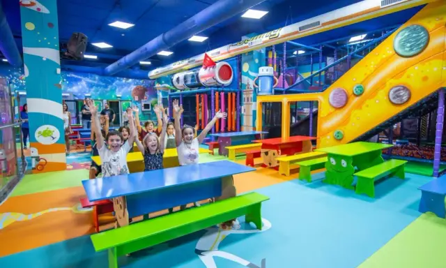 Un grupo de niños alegres levanta las manos en un vibrante centro comercial con juegos para niños, con toboganes, túneles y coloridos muebles.