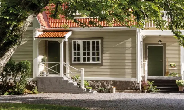 Ett charmigt hus med tegelrött tak och ljusgul fasad, redo att måla om huset. En stor ek skuggar entrén.