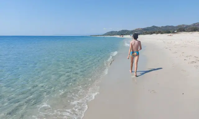 Un bambino cammina su una spiaggia di sabbia bianca, verso le spiagge più belle della Sardegna est. Acqua cristallina e cielo azzurro.