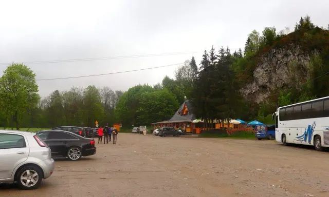 Parking przy Wąwozie Homole. Samochody, autokar i budynek zadaszony, otoczony drzewami, u podnóża skalnego zbocza.