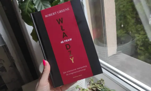 Dłoń z neonowym różowym lakierem trzyma książkę "48 praw władzy" Roberta Greene'a.